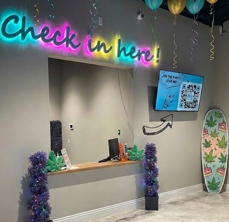 Reception desk with neon "Check in here!" sign, colorful balloons, and a surfboard with a leaf pattern.